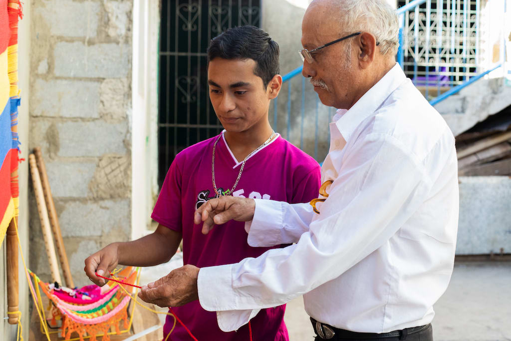 A older man teaches a teenage boy how to make a hammock.